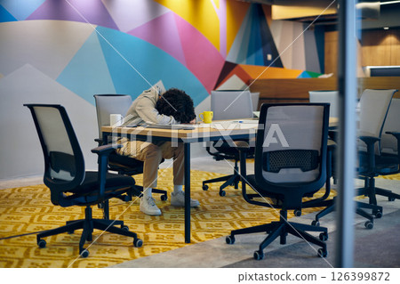 In a modern, spacious startup office, an exhausted African American man lies on a desk, reflecting the challenges of contemporary corporate life and the importance of work-life balance. 126399872