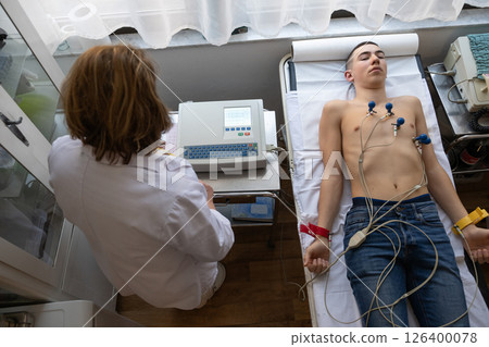 A female doctor performs an ECG on a young man in a hospital room, carefully monitoring his heart health during a routine checkup. A female doctor performs an ECG on a young man in a hospital room, carefully monitoring his heart health during a routine checkup. 126400078