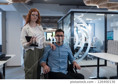 Young business colleagues, collaborative business colleagues, including a person in a wheelchair, walk past a modern glass office corridor, illustrating diversity, teamwork and empowerment in the Young business colleagues, collaborative business colleagues, including a person in a wheelchair, walk past a modern glass office corridor, illustrating diversity, teamwork and empowerment in the 126400136