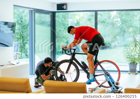 A cameraman filming an athlete riding a triathlon bike on a simulation machine in a modern living room. Training in pandemic conditions 126400255