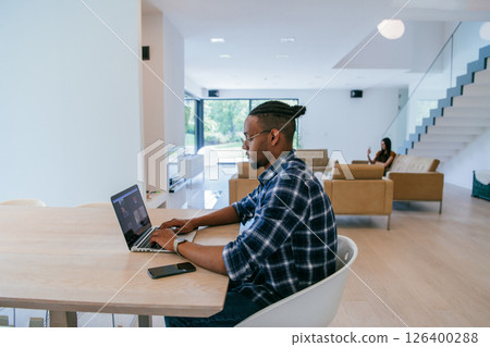 African American influencer in glasses sitting at a table in a modern living room, using a laptop for business video chat, conversation with friends and entertainment. 126400288
