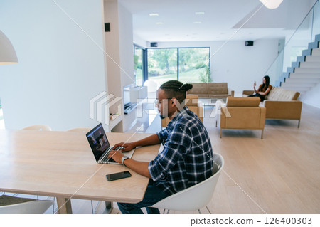 African American influencer in glasses sitting at a table in a modern living room, using a laptop for business video chat, conversation with friends and entertainment. 126400303