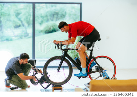 A cameraman filming an athlete riding a triathlon bike on a simulation machine in a modern living room. Training in pandemic conditions A cameraman filming an athlete riding a triathlon bike on a simulation machine in a modern living room. Training in pandemic conditions 126400341