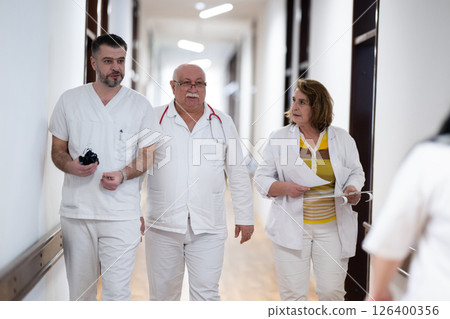 A group of healthcare professionals in white uniforms walking through a hospital hallway while reviewing patient documents and discussing treatment plans. A group of healthcare professionals in white uniforms walking through a hospital hallway while reviewing patient documents and discussing treatment plans. 126400356