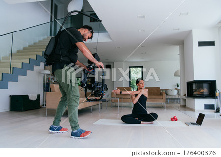 Behind the scene. Photo of a cameraman recording a woman doing yoga exercises at an online training while using and watching a laptop. Behind the scene. Photo of a cameraman recording a woman doing yoga exercises at an online training while using and watching a laptop. 126400376