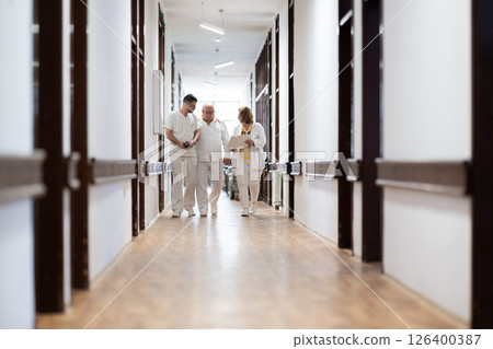 A group of healthcare professionals in white uniforms walking through a hospital hallway while reviewing patient documents and discussing treatment plans. 126400387