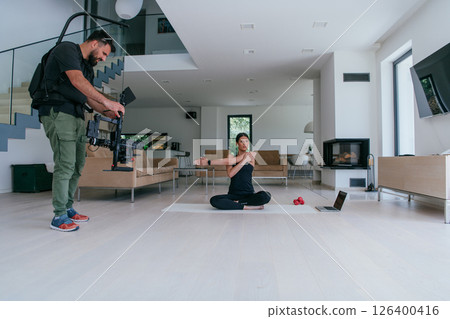 Behind the scene. Photo of a cameraman recording a woman doing yoga exercises at an online training while using and watching a laptop. 126400416