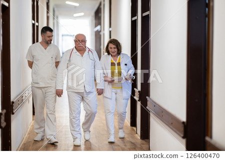 A group of healthcare professionals in white uniforms walking through a hospital hallway while reviewing patient documents and discussing treatment plans. A group of healthcare professionals in white uniforms walking through a hospital hallway while reviewing patient documents and discussing treatment plans. 126400470