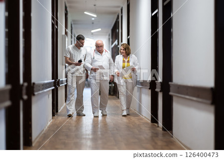 A group of healthcare professionals in white uniforms walking through a hospital hallway while reviewing patient documents and discussing treatment plans. 126400473