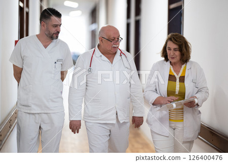 A group of healthcare professionals in white uniforms walking through a hospital hallway while reviewing patient documents and discussing treatment plans. A group of healthcare professionals in white uniforms walking through a hospital hallway while reviewing patient documents and discussing treatment plans. 126400476