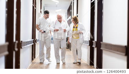 A group of healthcare professionals in white uniforms walking through a hospital hallway while reviewing patient documents and discussing treatment plans. 126400493