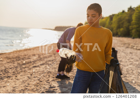 Earth day. Volunteers activists team collects garbage cleaning of beach coastal zone. Woman puts plastic bottle trash in garbage bag on ocean shore. Environmental conservation coastal zone cleaning Earth day. Volunteers activists team collects garbage cleaning of beach coastal zone. Woman puts plastic bottle trash in garbage bag on ocean shore. Environmental conservation coastal zone cleaning 126401208