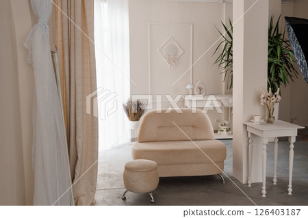 Elegant bridal room interior featuring a soft beige sofa, decorative wall accents, a white side table, and a wedding dress draped on a hanger, setting a romantic atmosphere 126403187