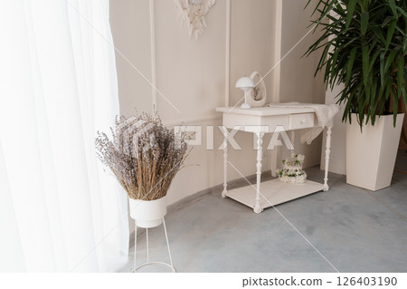 A serene interior scene with a delicate white console table adorned with lavender and potted plants against a neutral beige wall and sheer curtains 126403190