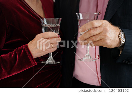 A sophisticated couple, a man and a woman, clink glasses of clear liquid in a dimly lit setting, suggesting celebration or a special occasion A sophisticated couple, a man and a woman, clink glasses of clear liquid in a dimly lit setting, suggesting celebration or a special occasion 126403192