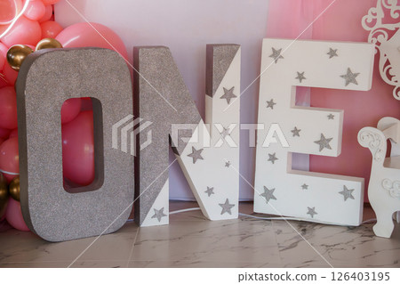 A glamorous first birthday celebration backdrop featuring large, sparkling silver "ONE" letter decorations adorned with stars, alongside a pastel pink balloon garland and a unicorn statue A glamorous first birthday celebration backdrop featuring large, sparkling silver "ONE" letter decorations adorned with stars, alongside a pastel pink balloon garland and a unicorn statue 126403195