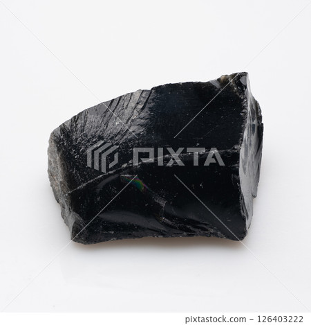 A polished, dark black obsidian rock with a slightly jagged surface and iridescent sheen resting on a plain white background, showcasing its glassy texture 126403222