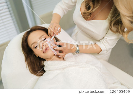 High-angle view of beautician applying pre-procedure markings on woman face using white pencil, preparing for cosmetic treatment in bright beauty salon. Concept of rejuvenating skin, preventing aging. 126405409