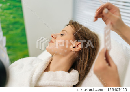 Beautician preparing patient facial skin for treatment in spa center, marking grid with white pencil. Cosmetologist applying pre-procedure markings on woman face, preparing for cosmetic treatment. 126405423