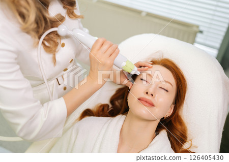 Top view of aesthetician using microneedling device performing rejuvenation procedure to female patient lying with closed eyes on treatment table, enhancing skin health and appearance in spa salon. 126405430