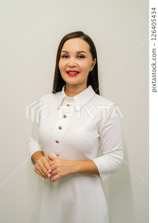 Professional female dermatologist wearing white coat and red lipstick, standing confidently on white isolated background, smiling looking at camera, radiating medical expertise and skincare knowledge. 126405434