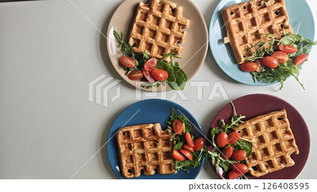 Four plates of golden waffles served with fresh cherry tomatoes and leafy greens on a white table. Concept of healthy breakfast, modern food styling, and colorful presentation 126408595