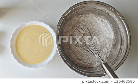 Bowl of flour and whisk next to a bowl of liquid waffle batter on a white surface. Concept of homemade cooking, preparation process, and baking simplicity 126408596