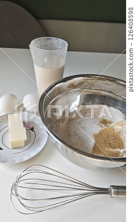 Baking ingredients on a table including flour, sugar, eggs, butter, milk, and a whisk in a clean kitchen setup. Concept of home baking, preparation process, and culinary creativity 126408598