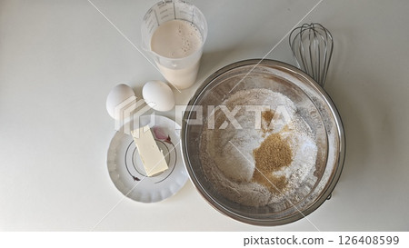 Flat lay of baking ingredients including flour, sugar, butter, eggs, milk, and a whisk on a white surface. Concept of home cooking, ingredient simplicity, and clean food preparation Flat lay of baking ingredients including flour, sugar, butter, eggs, milk, and a whisk on a white surface. Concept of home cooking, ingredient simplicity, and clean food preparation 126408599