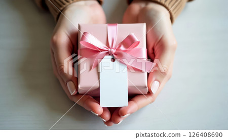 Female hands holding pink gift box with ribbon and blank tag on white wooden background. Celebration and giving. 126408690