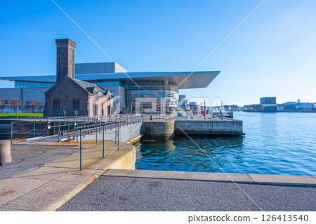 Visitors admire the architecture of Copenhagen Opera House on a clear sunny day, enjoying the waterfront view and vibrant atmosphere near the lively cityscape. 126413540