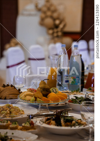 Lavishly Set Banquet Table with Fresh Fruit, Pastries and Beverages in Elegant Dining Room Interior, Featuring White Chair Covers and Sculptural Elements Lavishly Set Banquet Table with Fresh Fruit, Pastries and Beverages in Elegant Dining Room Interior, Featuring White Chair Covers and Sculptural Elements 126413890