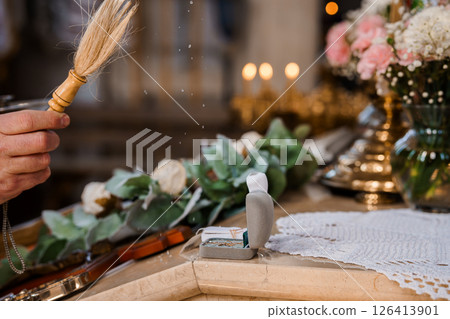 Orthodox wedding ceremony detail featuring a priest sprinkling holy water with a brush over rings and flowers on an altar adorned with candles and decorations Orthodox wedding ceremony detail featuring a priest sprinkling holy water with a brush over rings and flowers on an altar adorned with candles and decorations 126413901