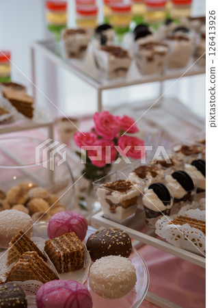 A beautifully arranged dessert table featuring colorful mini cupcakes, chocolate treats, macarons, and other pastries on transparent stands with pink roses 126413926