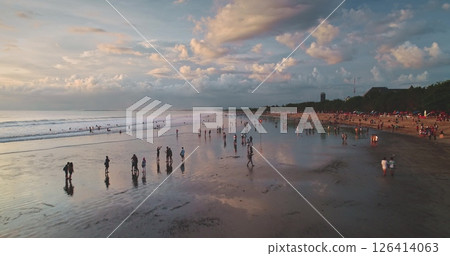 Tourists walking along the sandy shore at Dreamland Beach in Bali, Indonesia, are captured in an aerial drone view during a stunning sunset, surrounded by a dramatic, cloud-filled sky 126414063