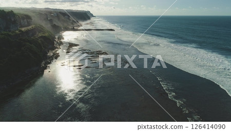 Aerial drone footage capturing the sun reflecting on the shallow water of a rocky coast in Bali, Indonesia, with gentle waves rolling in under a cloudy sky 126414090