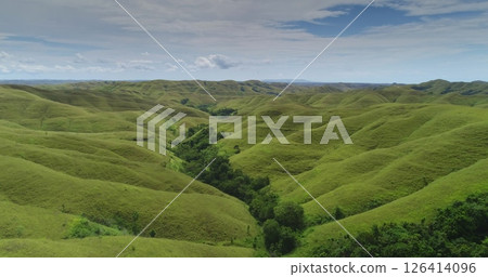 Rolling green hills extend as far as the eye can see across the landscape of Sumba island, Indonesia, with a line of trees growing along a valley and a partly cloudy sky overhead 126414096
