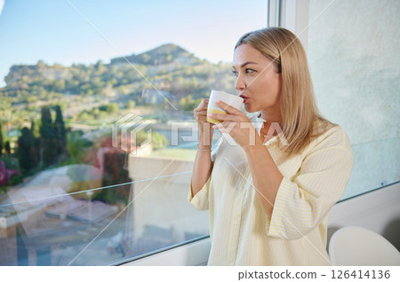 Woman in Cozy Home Enjoying Beverage with Serene Landscape Views 126414136