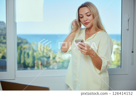 Casual Woman Using Smartphone by a Window with Scenic Ocean View 126414152