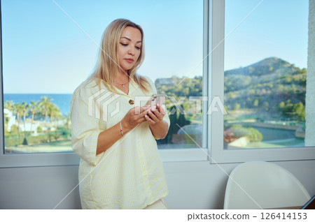 Woman Checking Her Phone in a Bright Room, Overlooking a Scenic Coastal View 126414153