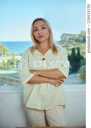 Confident Woman in a Bright Blouse Standing by a Window Overlooking a Scenic Natural Landscape Confident Woman in a Bright Blouse Standing by a Window Overlooking a Scenic Natural Landscape 126414156