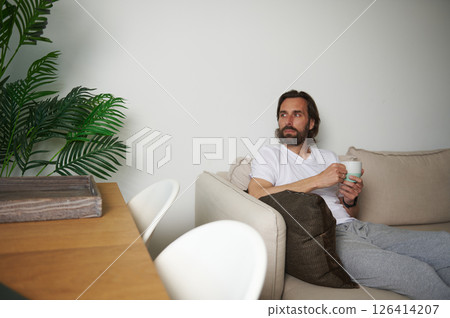 Man Relaxing on Sofa with Coffee in Cozy Minimalist Home Setting Man Relaxing on Sofa with Coffee in Cozy Minimalist Home Setting 126414207