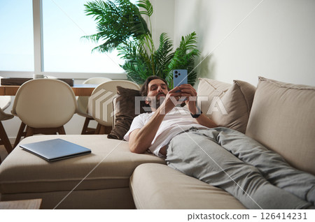 Man Relaxing on Couch Using Smartphone in a Comfortable Modern Living Room Man Relaxing on Couch Using Smartphone in a Comfortable Modern Living Room 126414231