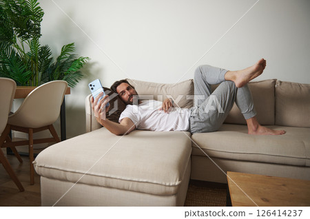 Man Relaxing on Sofa Holding Smartphone in Cozy Minimalist Living Room Setting Man Relaxing on Sofa Holding Smartphone in Cozy Minimalist Living Room Setting 126414237