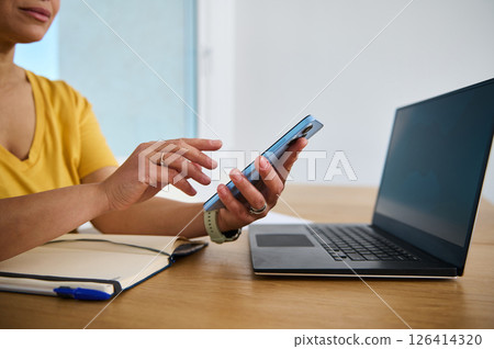 Person Using Smartphone and Laptop at Home Desk in a Bright Room 126414320