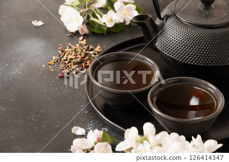 Black tea in cast iron teapot and cups decorated and white spring blossom flowers on black table. Chinese beverage. Tea ceremony. Close up. Copy space. 126414347