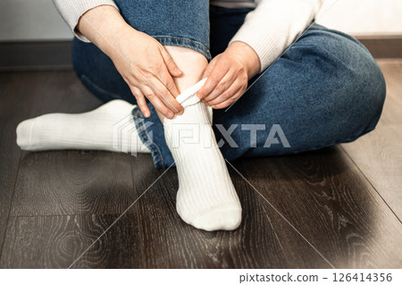 Relaxed moment as woman wears white socks, sitting on wooden floor. Natural lighting, perfect for comfort themes. 126414356