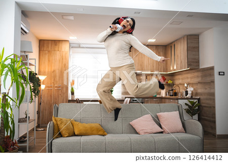 Woman with wireless headphones jumping on sofa and singing at home 126414412