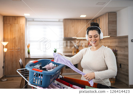 Woman listening to music while hanging the laundry on drying rack at home Woman listening to music while hanging the laundry on drying rack at home 126414423