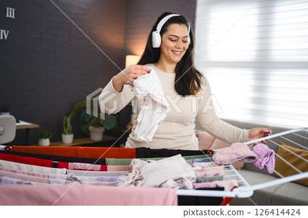 Woman listening to music while hanging the laundry on drying rack at home Woman listening to music while hanging the laundry on drying rack at home 126414424
